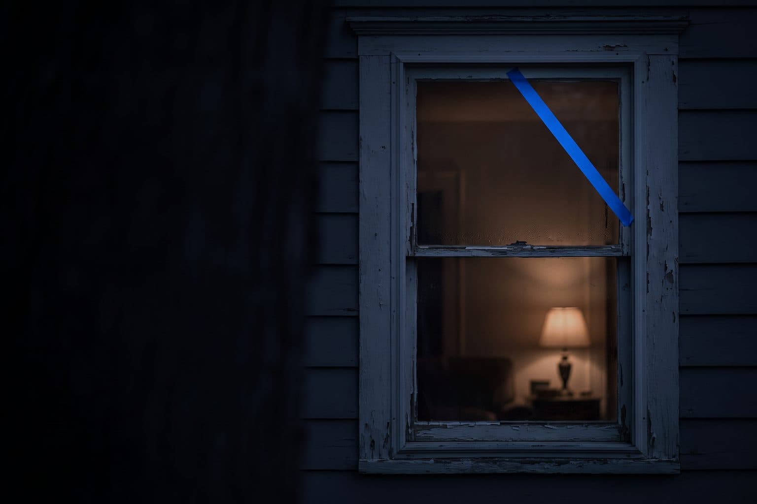 An old wooden sash window with peeling paint at night, blue tape across the glass