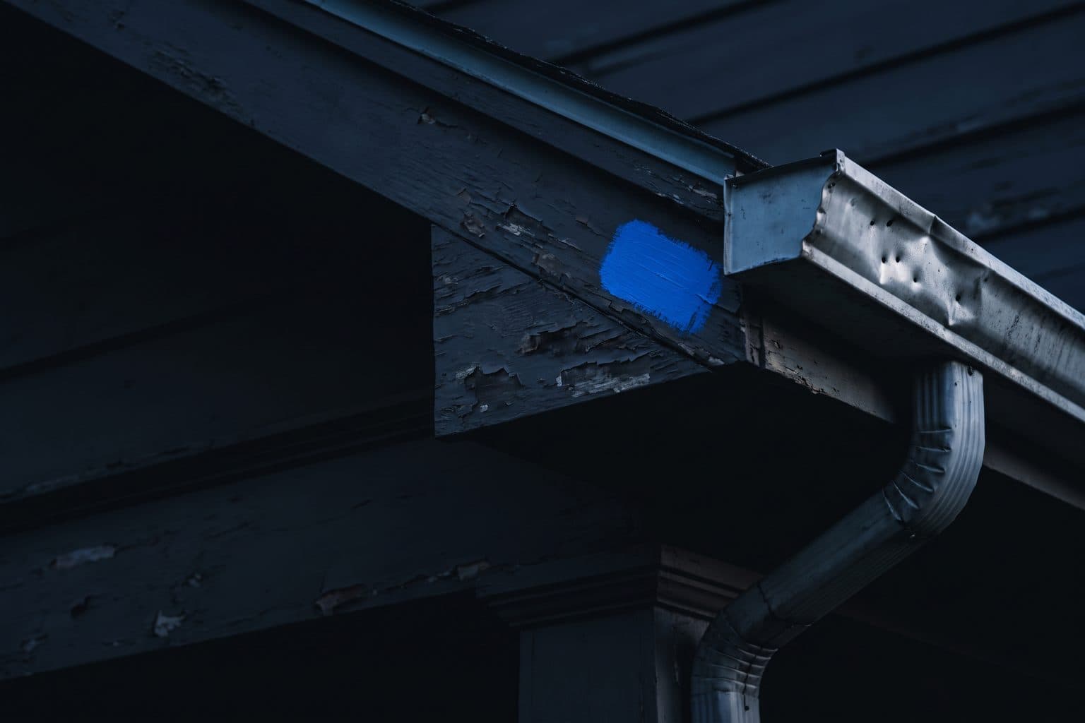 A hail-dented gutter and bent downspout under an eave with peeling paint, a fresh blue test swatch brushed on the trim